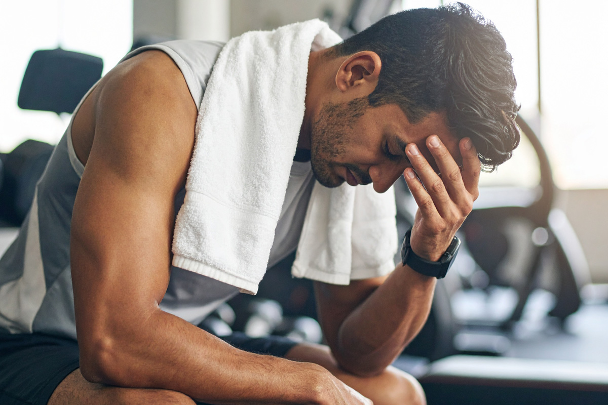 A visibly exhausted man sitting in a gym with his head in his hand and a towel around his neck, showing signs of dehydration after exercise.